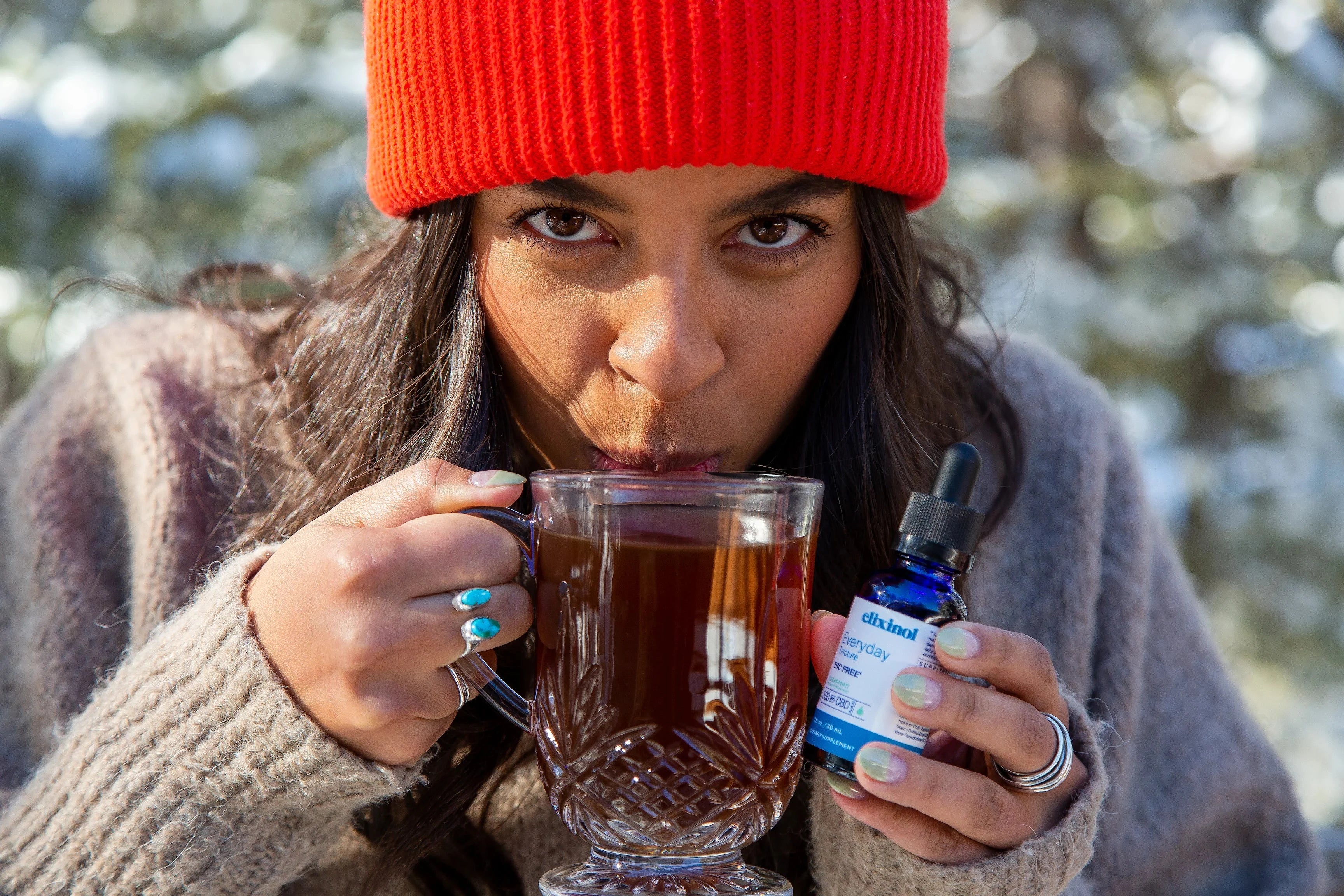A woman wearing a beanie outside sipping tea and holding an Elixinol tincture