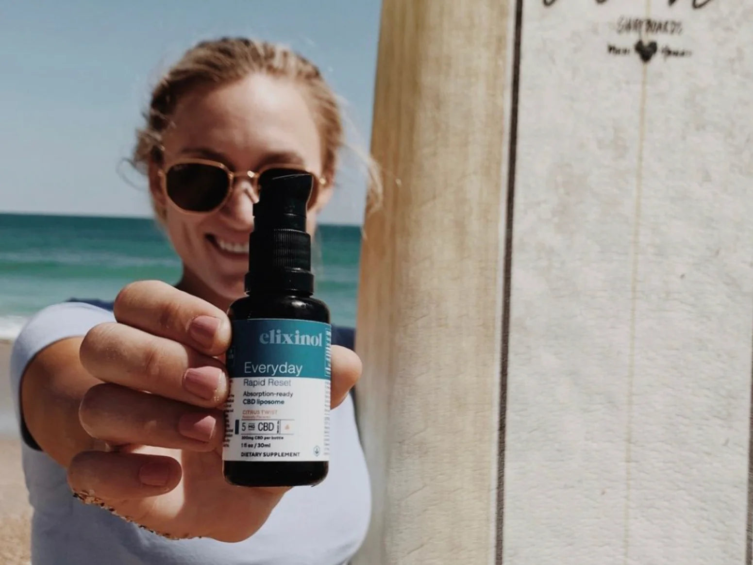 A woman smiling at a beach holding up a bottle of Elixinol Rapid Reset